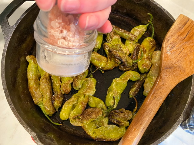 Shishito peppers that have been perfectly sautéed in a cast iron skillet with a wooden spoon resting on the pan and a salt grinder above the peppers