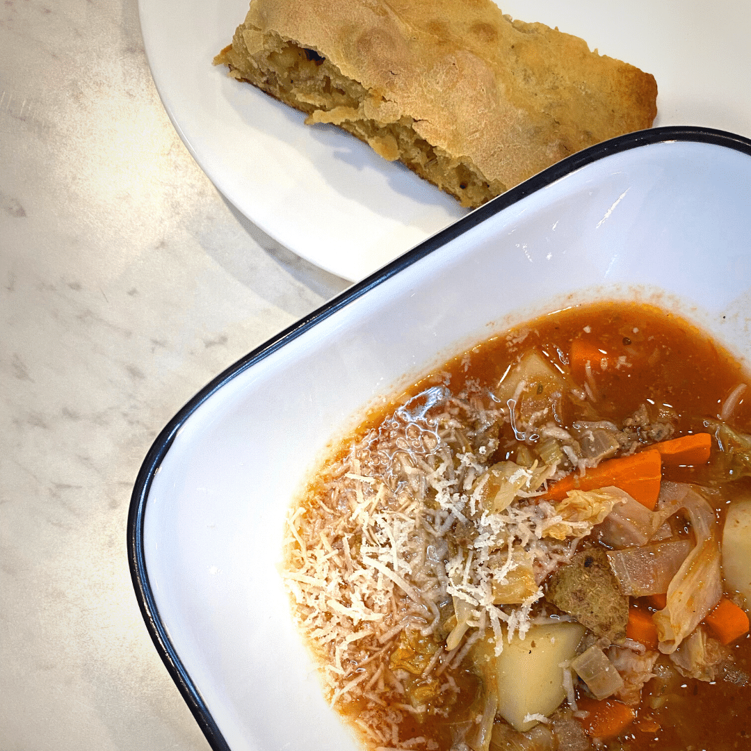 bowl of cabbage and beef soup in a white bowl next to a white plate with a piece of bread all atop a white counter