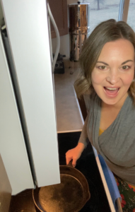 woman in a grey shirt standing by a stove with a cast iron skillet