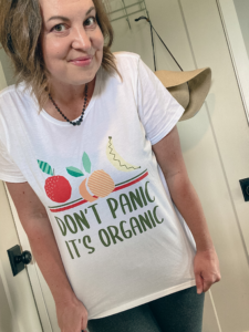 woman standing in front of a door with a sun hat hanging from a hook, and her shirt reads: don't panic its organic