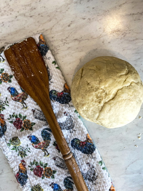 A towel with chickens, a wooden spoon, and a round dough ball for sourdough discard tortillas sit on a white counter.