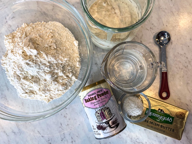 Four glass containers are sitting on a white counter along with baking powder, butter, and a measuring spoon ready to mix sourdough discard tortillas.