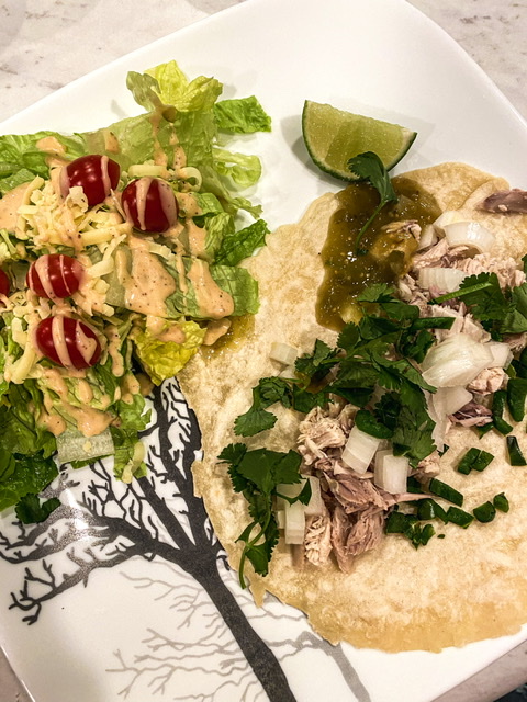 A sourdough discard tortilla is shown on a white plate topped with chicken, onions, cilantro, salsa verde which is next to a side salad topped with tomatoes and a white dressing with a lime wedge on the side.