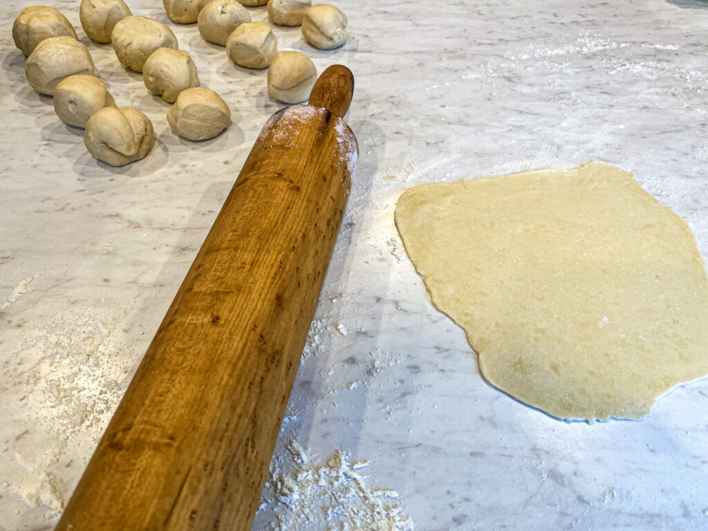 Sourdough discard tortilla dough balls are sitting on a white counter beside a wooden rolling pin and a flat tortilla.