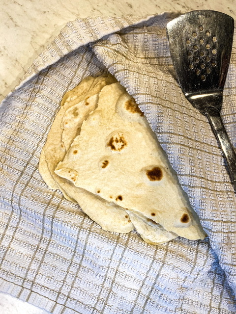 Sourdough discard tortillas are shown sitting inside a towel with grey stripes beside a metal spatula.