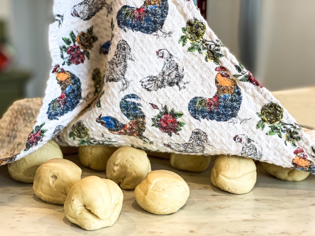 A towel with chickens is shown lifting off of small sourdough discard tortilla balls resting on a white counter.