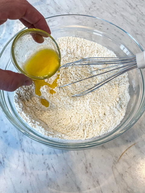 A glass bowl filled with flour sits on a white counter top while a small bowl of melted butter is poured into the flour to create sourdough discard tortillas. 