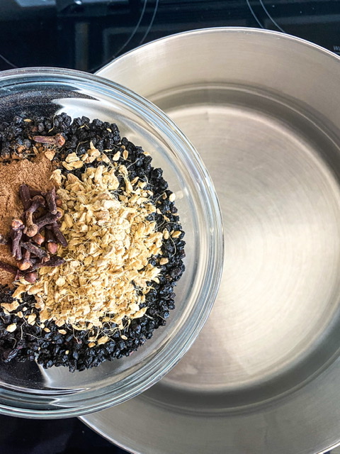 dried elderberries, ginger, cinnamon, and cloves in a glass bowl held over a stock pot of water