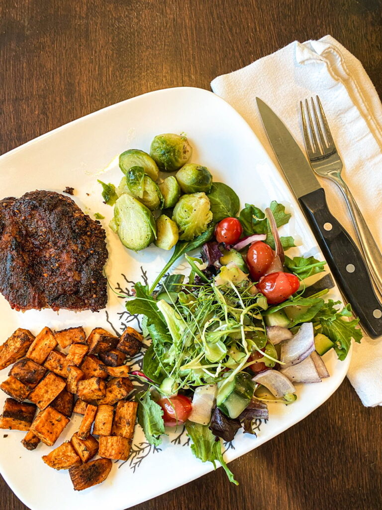 white plate filled with savory sweet potatoes, Brussels sprouts, salad, and pork steak