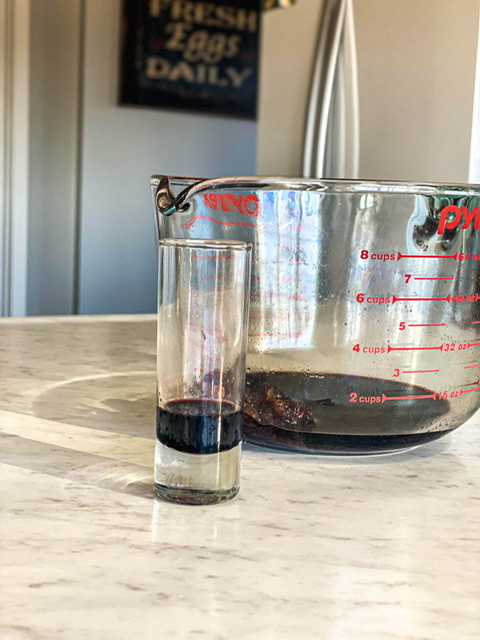 elderberry syrup in a glass measuring cup sitting next to a shot gloss of elderberry syrup on a white counter