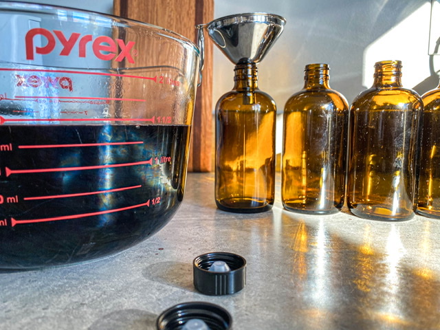 amber jars sitting on a grey counter with a stainless funnel next to a glass measuring cup filled with elderberry syrup