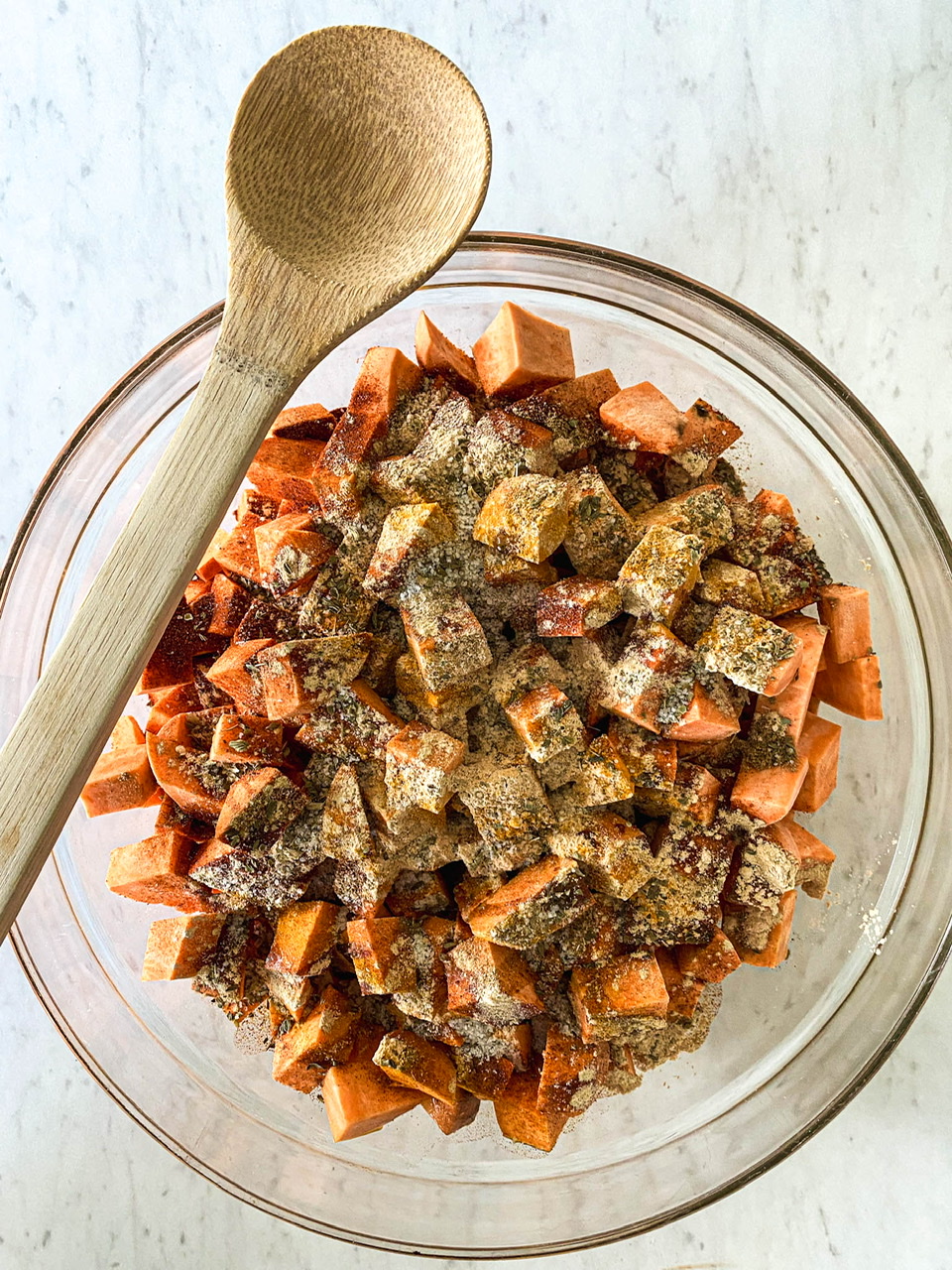 glass bowl on a white counter with a wooden spoon on the edge and filled with diced savory sweet potatoes