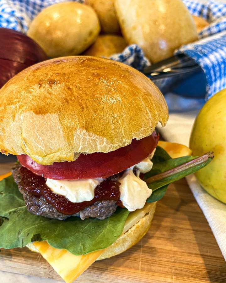 apple cheddar beef burger on a wooden cutting board with toppings and a bun