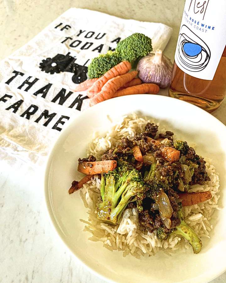 beef and broccoli stir-fry over a bed of rice in a white bowl next to raw ingredients to make the recipe and a bottle of wine