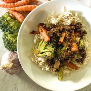 beef and broccoli stir-fry over a bed of rice in a white bowl next to raw ingredients to make the recipe