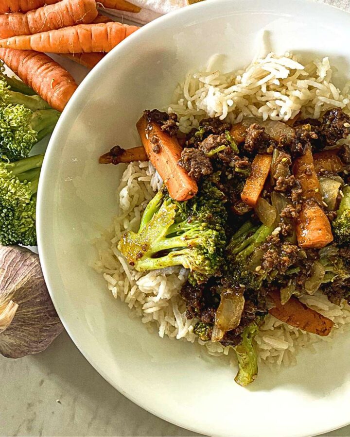 beef and broccoli stir-fry over a bed of rice in a white bowl next to raw ingredients to make the recipe